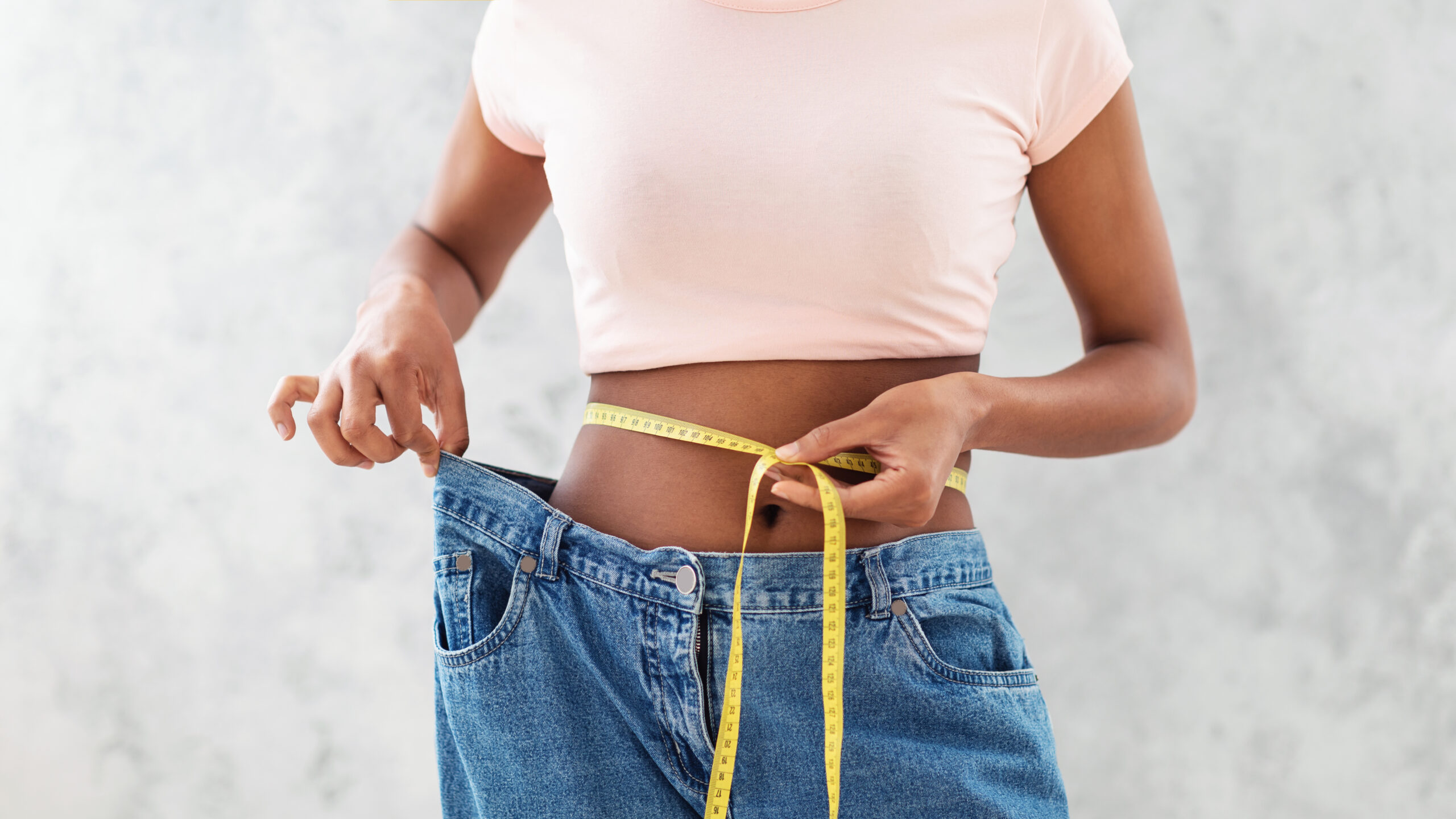 Black woman in old big jeans measuring her waist, showing results of slimming diet or liposuction